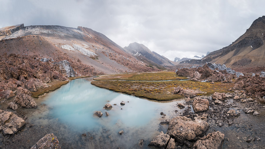 A milky turquoise tarn pooled among red-brown rock in the upper Brazeau canyon, meadow and distant peaks under grey sky
