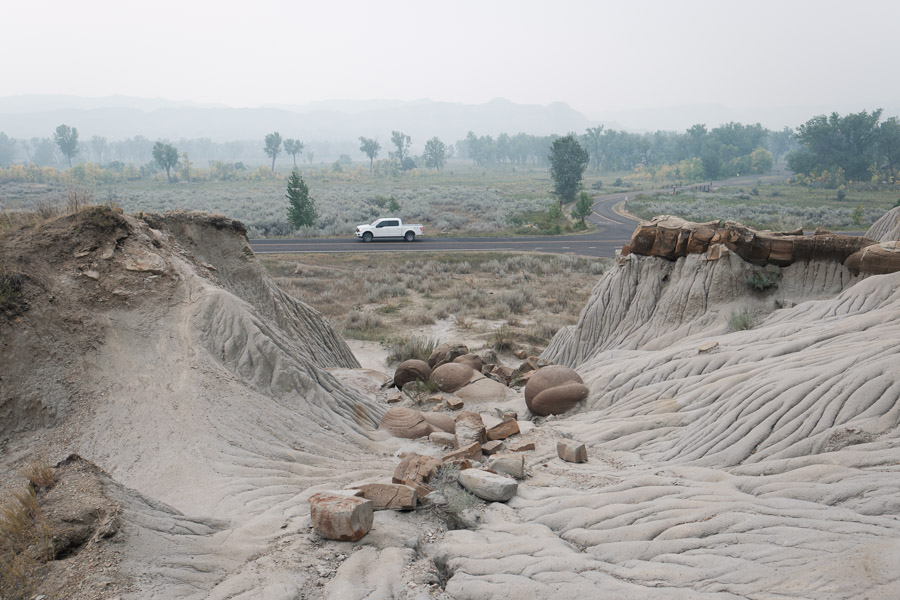 Round cannonball concretions weathering out of rippled grey badlands beside a road where a white pickup passes in wildfire haze