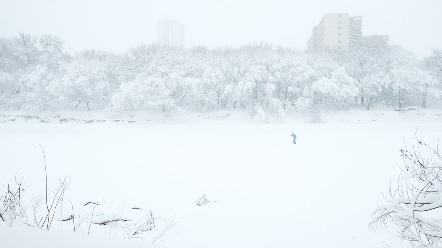 A lone cross-country skier crossing the frozen river in a whiteout, snow-laden trees and apartment towers dissolving into white behind