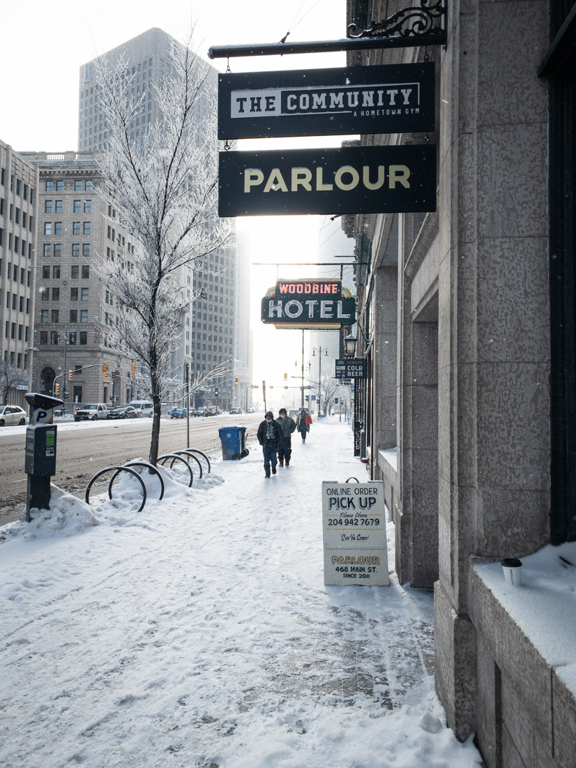 A snowy downtown Winnipeg sidewalk with pedestrians walking beneath the Woodbine Hotel's neon sign and frosted trees