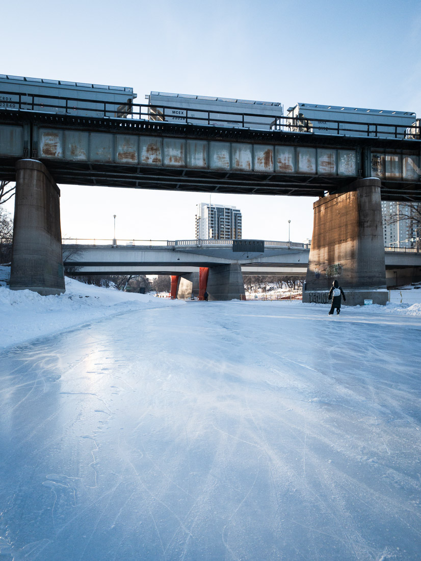 A skater glides along the river skating trail beneath a rusted rail bridge, fresh ice gleaming in the winter sun
