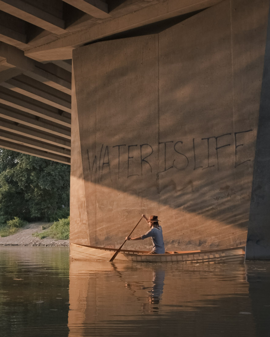 A canoeist paddles beneath a concrete Winnipeg bridge pillar marked with large graffiti reading WATER IS LIFE