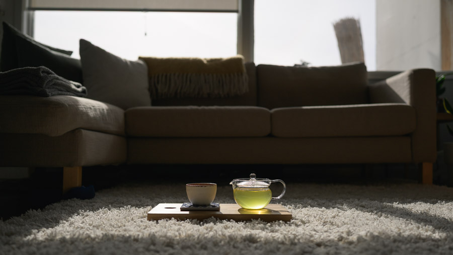 A glass teapot of green tea and a small ceramic cup on a wooden board, glowing in a beam of sunlight on a living room rug
