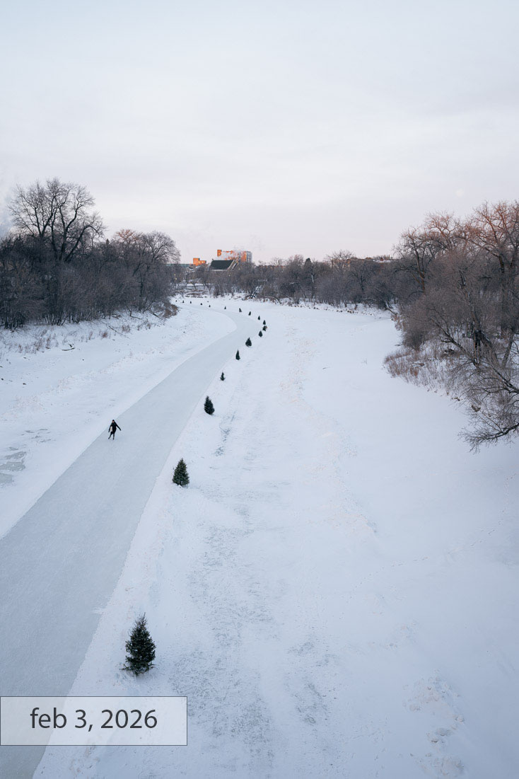 Labeled feb 3, 2026: a lone skater glides down the river skating trail at dusk, the ice lane lined with small evergreen trees