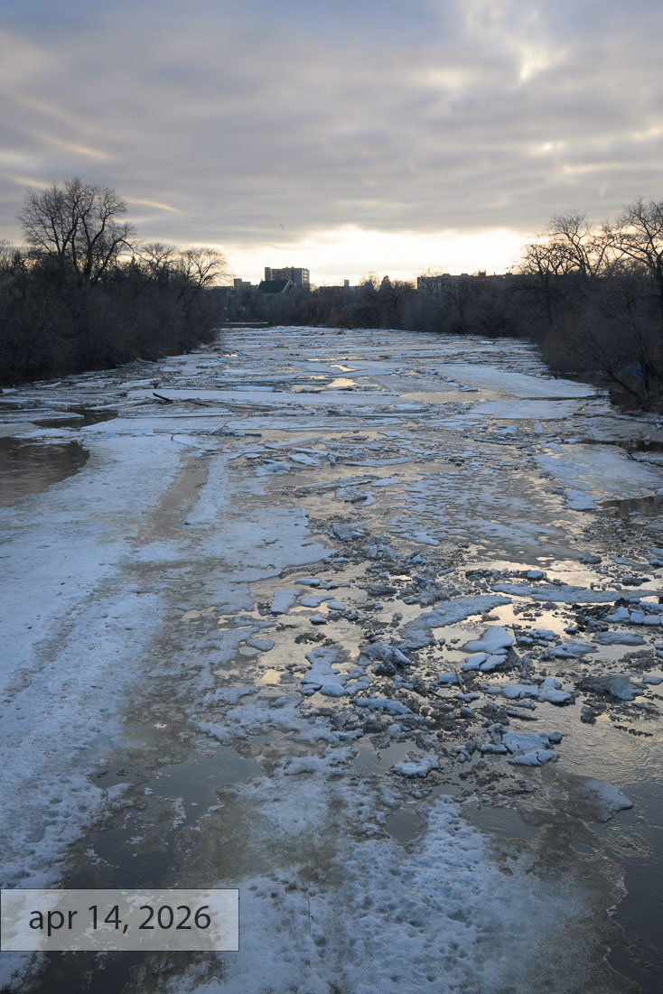 Labeled apr 14, 2026: broken ice floes drifting on the darkening river at sunset, reflecting a moody sky