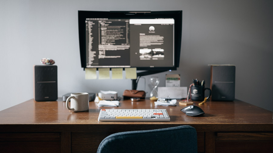 A wooden desk with a wall-mounted monitor showing a code editor beside a web page, sticky notes along its lower edge, a mechanical keyboard, mouse, ceramic mug, and bookshelf speakers